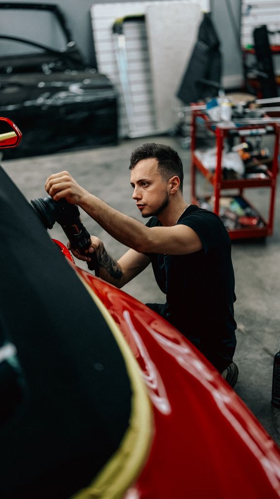 A focused young mechanic carefully polishes a red car in an automotive workshop setting.